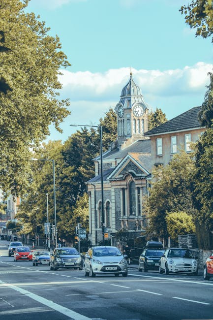 A view of Anerley Road in Crofton Park during daytime, showing parked cars lining the street and a historic clock tower building with a domed roof and spire in the background. The street is shaded by tall trees with green foliage, and the sky is partly cloudy with patches of blue. The scene depicts an urban environment suitable for residential, house removal, or furniture transport activities, with visible pavement, road markings, and street lighting. The image captures a typical setting for home relocation processes and the logistics involved in loading or unloading furniture and boxes in a residential area, consistent with professional removals services provided by Man with Van Crofton Park.