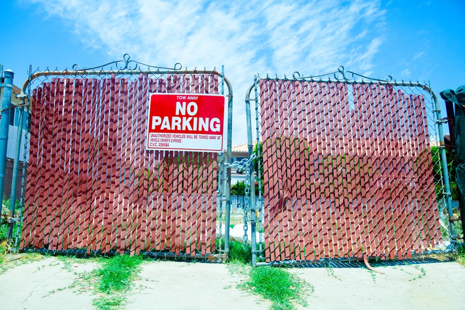 A metal gate with a chain latch, painted in dark colors, blocking entrance to a property, with a prominent red and white 'No Parking' sign attached to the left side of the gate. The sign indicates towing away for unauthorized vehicles, and the gate is mounted on blue metal posts. Behind the gate, there is a glimpse of a narrow driveway with a small area of grass and vegetation on the pavement in front. The sky above is bright and clear with some light cloud cover. This scene likely depicts a parking restriction area adjacent to a residential or commercial property, relevant to the logistics of house removals and moving services provided by Man with Van Crofton Park, who manage efficient access for furniture transport and loading during a home relocation.