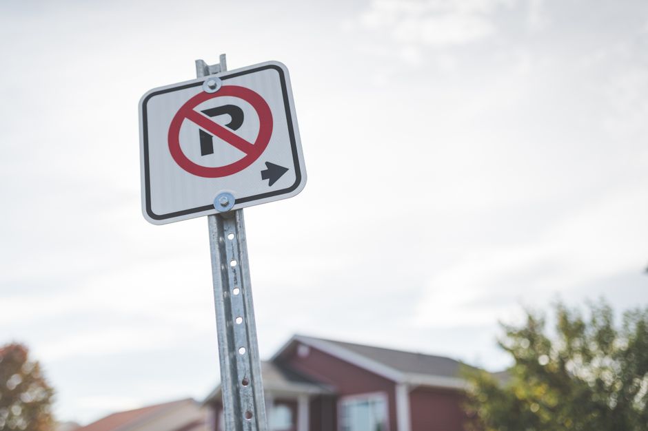 A rectangular white traffic sign mounted on a metal pole displaying a black letter 'P' with a red circle and slash through it, indicating no parking, with a black arrow pointing to the right located in an outdoor setting. In the background, there are residential houses with sloped roofs, partly blurred trees, and a cloudy sky, suggesting a suburban street environment. The sign is positioned in the foreground, capturing the context of parking regulations relevant to house removals and loading areas; the image emphasizes the importance of understanding local parking and loading bay rules during home relocation or furniture transport, with the sign potentially indicating restricted parking zones near residential properties where professional moving services, such as those provided by Man with Van Crofton Park, operate.
