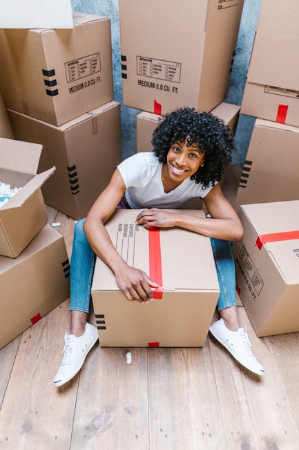 A man and woman inside a room filled with cardboard boxes during a home relocation process. The man, wearing a grey hoodie, sits on the floor with his arm resting on a box, looking attentively at the