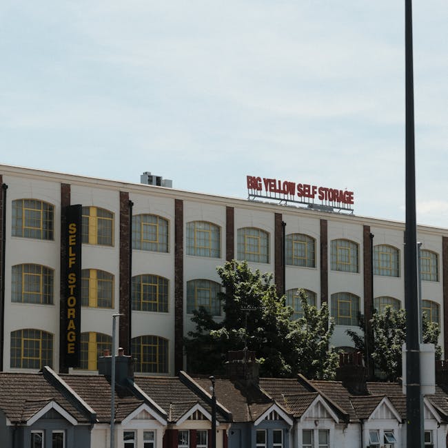 The image shows the exterior of a large, multi-story self-storage facility with white walls and multiple rows of tall, rectangular windows framed in yellow. The building features a prominent vertical yellow sign on the left that reads 'SELF STORAGE,' and a red sign on the roof that states 'BIG YELLOW SELF STORAGE.' In front of the building, there are small residential houses with dark tiled roofs, chimney stacks, and white window frames, partially obscured by green trees and shrubs. The scene is captured during daytime with a partly cloudy sky overhead and a lamppost visible in the foreground, indicating an urban setting typical for house removals and furniture transport logistics, as referenced in the page titled 'Bulky-item Removals in SE4: Costs & Council Rules' from manwithvancroftonpark.co.uk.
