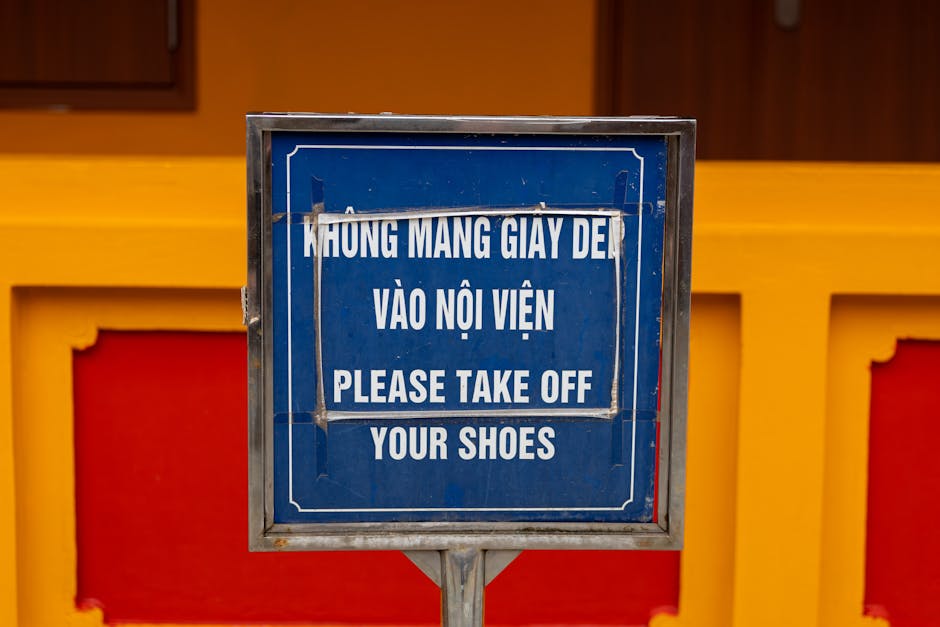 A blue sign with white text mounted on a metal stand, displaying a message in Vietnamese and English that reads 'HƯNG MANG GIÀY ĐẾ VÀO NỘI VIỆN PLEASE TAKE OFF YOUR SHOES,' placed in front of a decorative wall featuring yellow and red panels. The sign indicates an area where visitors are required to remove their shoes before entering, likely within a residential or indoor communal space. The surrounding environment suggests it is positioned at the entrance of a property, possibly during a house removal or relocation process. The presence of the sign and the colorful background provide context for a home environment, with the signage acting as a prompt for guests and movers involved in furniture transport or packing and moving activities, aligning with the services provided by Man with Van Crofton Park.