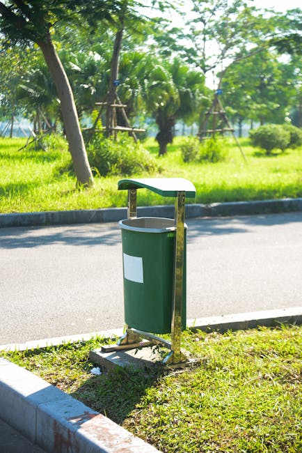 A green outdoor public rubbish bin with a white blank label, mounted on a metal stand attached to a concrete curb, situated beside a paved road. Behind the bin, there is a grassy area with trees, including palm trees, supported by wooden stakes, under bright daylight with clear skies. The scene suggests a park or suburban street environment, with no visible moving or relocation-related objects or activity.