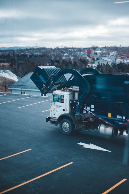 A white commercial rubbish collection truck with a black waste container attached is parked on a nearly empty rooftop parking lot during daytime, with yellow parking lines visible on the asphalt surface. The truck is equipped with hydraulic arms and lifting mechanisms for waste disposal. In the background, there are residential and industrial buildings with rooftops, trees, and rolling hills under a cloudy sky. The scene reflects an urban area engaged in waste collection or commercial refuse removal, which could be part of logistics involved in house removals or move-related services offered by Man with Van Crofton Park. The image highlights the vehicle's positioning on the rooftop, ready for waste collection or disposal as part of a home relocation or furniture transport process.