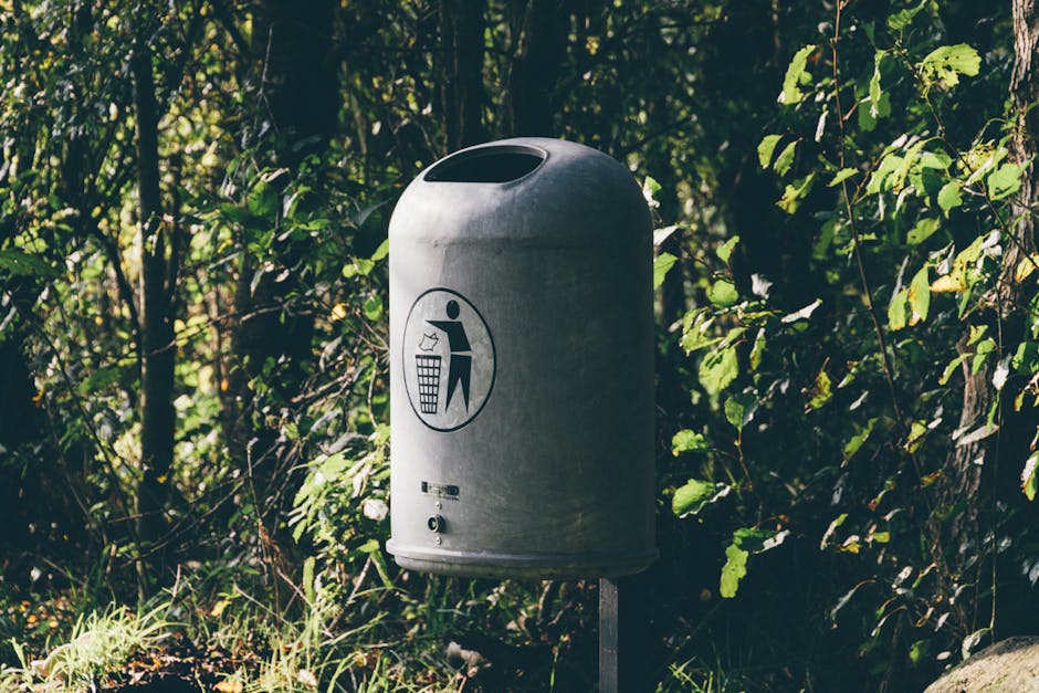 A green outdoor public rubbish bin with a white blank label, mounted on a metal stand attached to a concrete curb, situated beside a paved road. Behind the bin, there is a grassy area with trees, including palm trees, supported by wooden stakes, under bright daylight with clear skies. The scene suggests a park or suburban street environment, with no visible moving or relocation-related objects or activity.