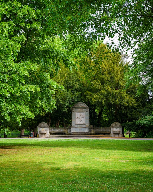 A stone monument situated in a park area is partially visible behind dense green foliage from mature trees with bright green leaves. The monument appears to be made of light-colored stone and features a central rectangular plaque with a carved relief or inscription, flanked by two smaller stone structures that resemble planters or decorative elements. The grassy area in the foreground is well-maintained, with short, lush grass, and the scene is illuminated by natural daylight, suggesting a clear or partly cloudy day. The park environment, with its mature trees and neatly kept lawn, provides a peaceful setting typical of a city park or public green space, where residents might gather or go for leisure activities. This outdoor scene complements the context of a professional house removal or relocation service offered by Man with Van Crofton Park, illustrating a typical environment where moving companies may operate when transporting furniture or packing materials from or to a residential property.