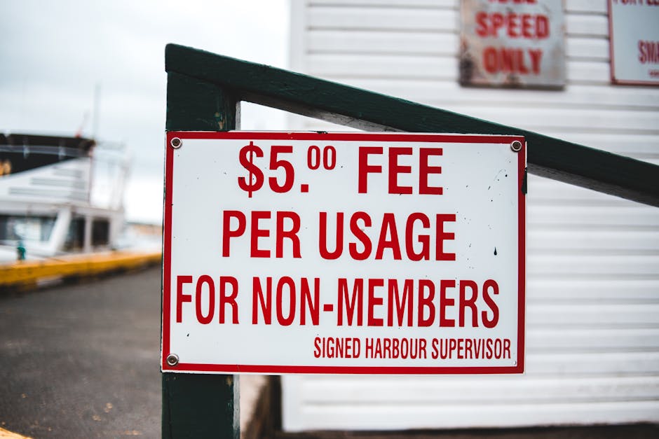 Close-up of a rectangular white sign with red text attached to a green metal post near a harbour area. The sign displays a fee of five pounds per usage for non-members, indicating a charge for accessing certain facilities or services, signed by the harbour supervisor. In the background, there are portions of boats docked at the harbour, with some visible mooring lines and parts of a dock or pier made of dark asphalt. A white building or shed with visible signage is partially seen behind the sign, and the overall environment is outdoor with natural daylight. The sign is part of a moving or logistics setting at a harbour, emphasizing the importance of fee regulation and access control related to property or service use, with the focus on safety and regulatory compliance typical of a relocation context involving maritime transport or storage areas.
