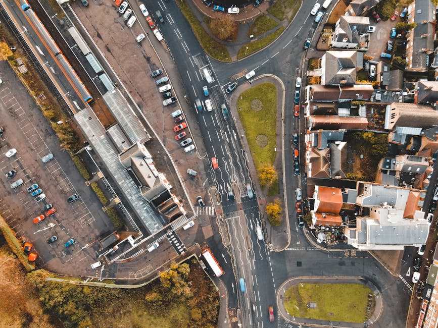 An aerial view of a busy urban intersection showing multiple lanes of traffic with cars moving in different directions, including small passenger vehicles and buses. Surrounding the roads are various buildings, including a large structure with a glass roof section, residential houses with brick facades, pitched roofs, and small gardens. On the pavement, a man from Man with Van Crofton Park, a professional removals company, is seen loading furniture wrapped in plastic and blankets into a large white van parked at the curb. The van’s sliding door is open, revealing interior packing materials like cardboard boxes, packing paper, and bubble wrap. Nearby, a trolley is positioned to assist with the transportation of packed items, and the area is well-lit with natural daylight. This scene captures the logistics involved in home relocation, including furniture transport, packing preparations, and the loading process during a house move near Crofton Park Station, as part of professional removals services.
