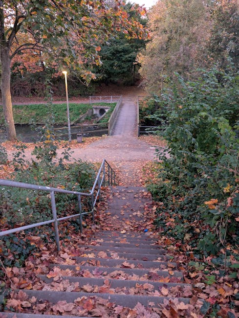 A set of outdoor concrete stairs with eleven steps, situated between green foliage on both sides, leading up to a doorway or pathway at the top. The stairs are bordered by metal handrails on each side, which are painted blue and extend from the bottom to the top of the staircase. The steps appear weathered, with some dirt and debris visible on their surfaces. Surrounding the stairs are lush bushes and trees, providing a natural environment. This scene depicts an exterior property access point, common in residential areas, where house removals and moving services may assist with navigating narrow staircases in the Crofton Park area. The lighting suggests daytime, with sunlight filtering through the leaves, highlighting the greenery and the textured surface of the stairs, emphasizing the typical features encountered during home relocation or furniture transport processes.
