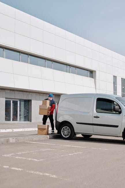 A person wearing a blue beanie, red jacket, and dark trousers is seen standing next to a silver cargo van, holding and stacking cardboard boxes during a home relocation. Several cardboard boxes are positioned on the ground near the van, with some being carried toward the vehicle. The van is parked on a paved surface in front of a modern white building with large windows and a minimalist façade. Inside the property, visible through the glass doors, there are also moving boxes, indicating active packing or unpacking processes. The scene captures the loading process typical of furniture transport and packing and moving activities carried out by Man with Van Crofton Park as part of a same-day, last-minute removals service, illustrating efficient logistics involved in residential relocations.