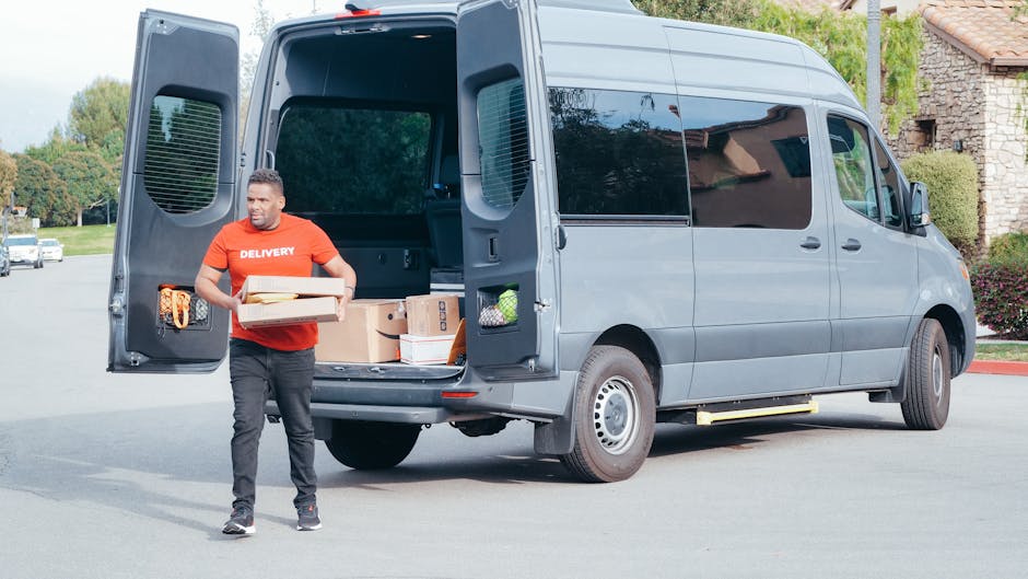 A man wearing a red delivery T-shirt is standing outside a grey van with its rear doors open, loading and unloading cardboard boxes and packing materials used for a home relocation. The interior of the van shows several stacked boxes, some wrapped in plastic and others in plain cardboard, along with packing paper and small items. The scene is set on a paved driveway with a residential building in the background, surrounded by greenery. The man is carrying a box while other boxes are placed inside the van, which is parked adjacent to a house with stone walls and a tiled roof. Trolleys, straps, and blankets are visible around the vehicle, indicating a professional furniture transport and moving process by Man with Van Crofton Park, experienced in last-minute removals and packing and moving services.