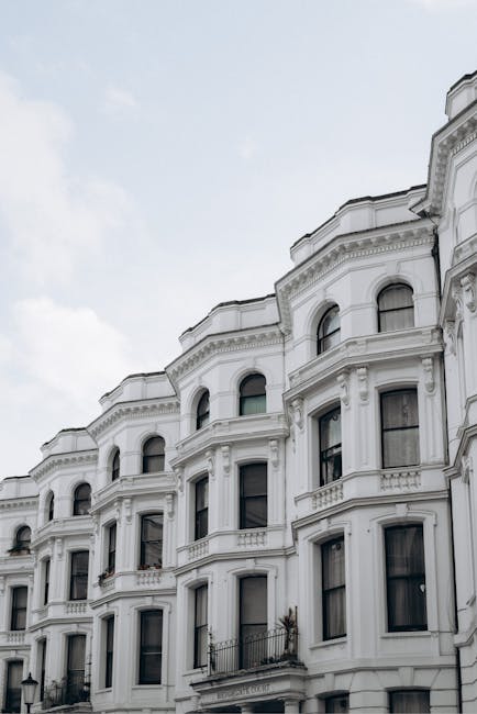 Photograph of a row of white Victorian terraced houses with ornate architectural details, including decorative cornices and arched windows, situated on Crofton Park. The image shows the exterior facades from street level, with some windows open and small balconies visible. The sky is overcast, providing diffuse lighting that highlights the building's features. In the foreground, part of a pavement and a black metal fence can be seen. This setting reflects a typical residential area suitable for home relocation services, such as those offered by Man with Van Crofton Park, which specialise in furniture transport, packing, and loading processes for house removals in the SE4 area.