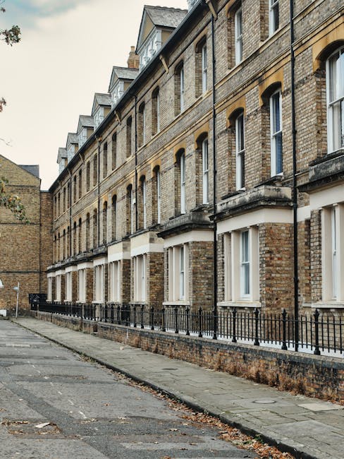 A row of traditional Victorian terraced houses constructed with yellowish-brown brickwork, featuring sash windows with white frames and decorative stone lintels. The buildings have small front gardens enclosed by black metal railings, with a narrow pavement running alongside the properties. An overcast sky provides diffuse natural light, and the street appears quiet with no visible pedestrians or vehicles. This setting exemplifies typical residential architecture found in Crofton Park, suitable for home relocation and furniture transport services by companies like Man with Van Crofton Park during moving and packing processes.